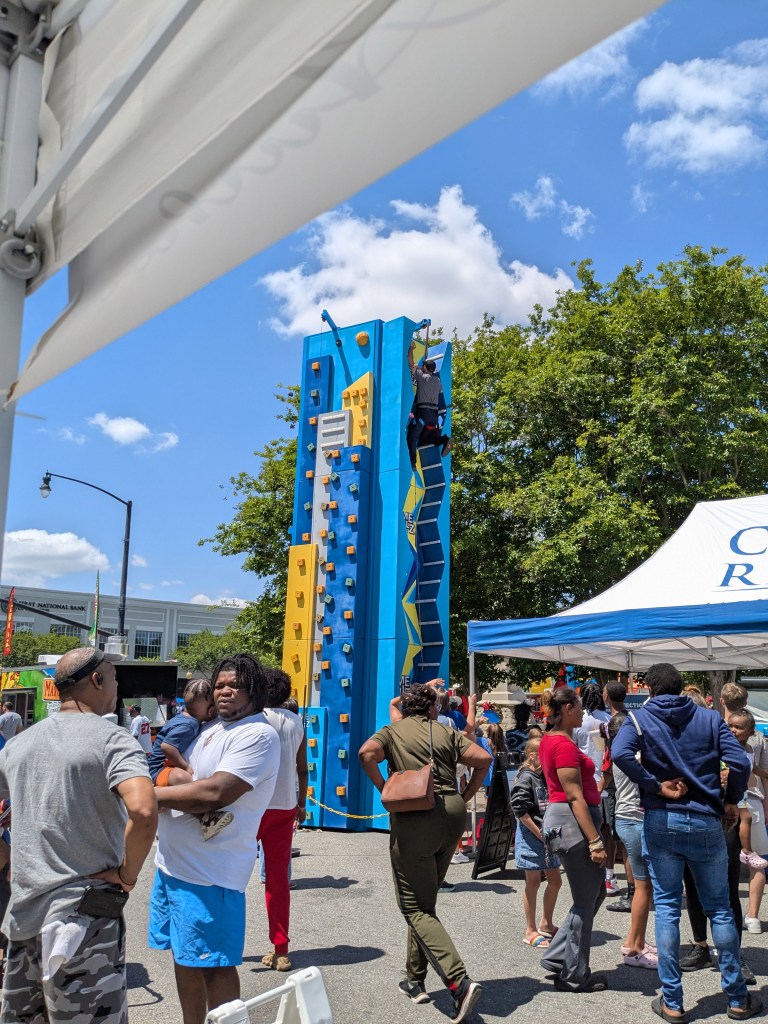 A climbing wall at a festival with a person climbing on it, surrounded by a crowd of people and colorful tents under a blue sky.